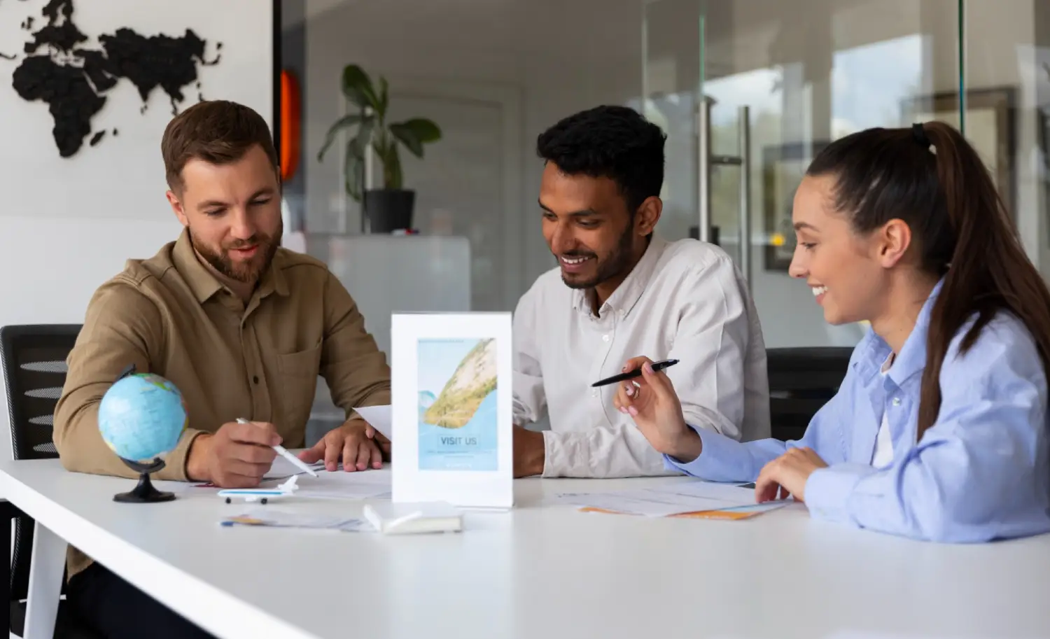 Three individuals collaborate at a modern conference table, discussing documents and ideas, with a small globe and promotional material nearby